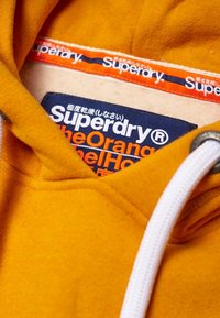 Sudadera de forro polar amarilla con un cordón blanco, que presenta una etiqueta azul y naranja con "Superdry" y "la etiqueta naranja".