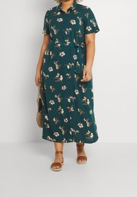 Teal bloemenjurk met knopen, korte mouwen en een strikceintuur. Heeft witte en oranje bloemenpatronen. Draagt met bruine sandaaltjes met banden.