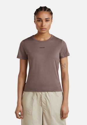 Femme aux cheveux tressés portant un t-shirt marron ajusté Icebreaker et un pantalon beige clair, posant devant un fond uni.