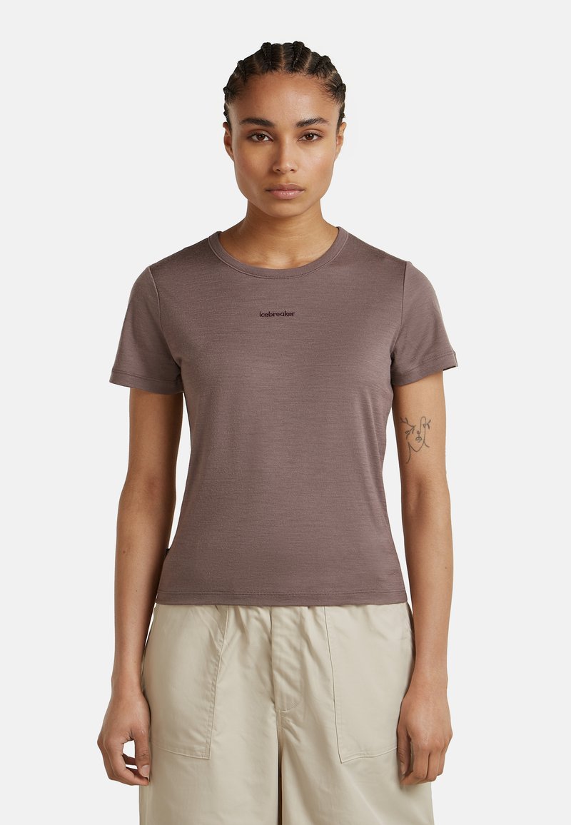 Frau mit geflochtenem Haar, die ein eng anliegendes braunes Icebreaker-T-Shirt und hellbeige Hosen trägt, steht vor einem schlichten Hintergrund.