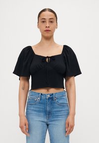 Svart cropped topp med puffermar, firkantet halslinje og en frontknute. Kombinert med lyseblå høytlivsjeans. Glatt stofftekstur.