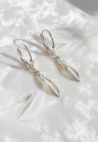 Silverörhängen med droppform och förlängd, bladliknande design; med en central rad av små klara ädelstenar. Bakgrunden har mjuka vita fjädrar.