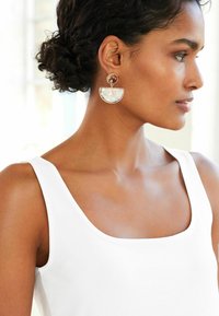 Sötét, göndör hajú nő, aki összefogta a haját, fehér ujjatlan felsőt visel és nagy arany-fehér félkör alakú lógó fülbevalót a jobb fülén, jobbra néz.