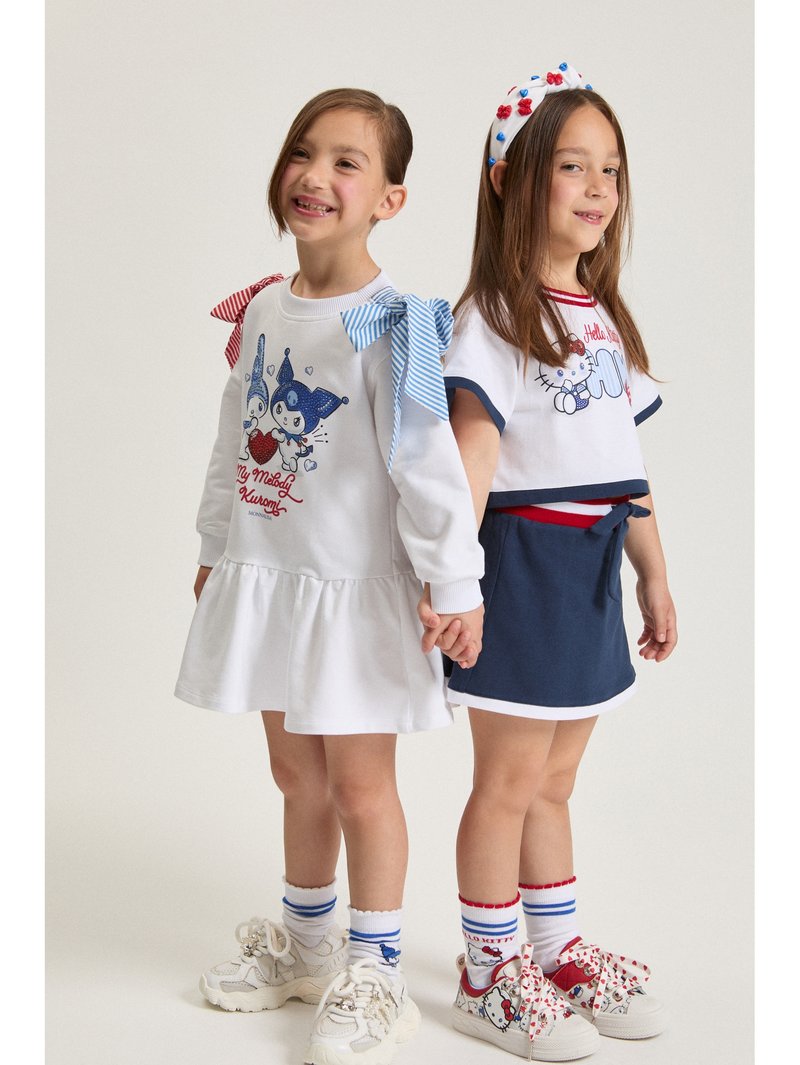 Dve dekleti, ki se držita za roke in nosita bele in mornarsko modre obleke s risanimi liki, ujemajoče se nogavice in superge, nasmejani v studijskem okolju.