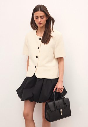 Krém színű, rövid ujjú kardigán fekete gombokkal, fekete, bő szoknyával párosítva. A modell egy fekete, struktúrált kézitáskát tart. Sima textúra.