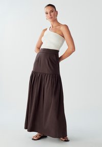 Hnedá, vrstvená maxi sukňa s priliehajúcim pásom; skombinovaná s bielym, rebrovaným topom na jedno rameno. Hedvábna látka a uvoľnený strih.
