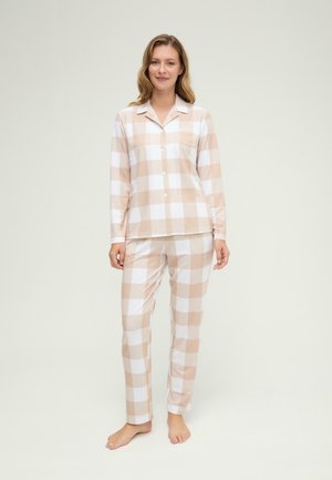 Mujer de pie descalza con pijama de pantalón y camisa de manga larga abotonada a cuadros beige y blanca sobre fondo liso.