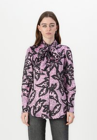 Bluza s dugim rukavima od ružičaste tkanine s crnim uzorkom lišća, mašna na ovratniku i dugmad na prednjoj strani. Glatka tekstura.