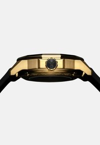 Goldfarbene Uhr mit sechseckigem Gehäuse, schwarzem Gummiarmband, strukturierten Akzenten und einer schwarzen Krone mit einem Logo.