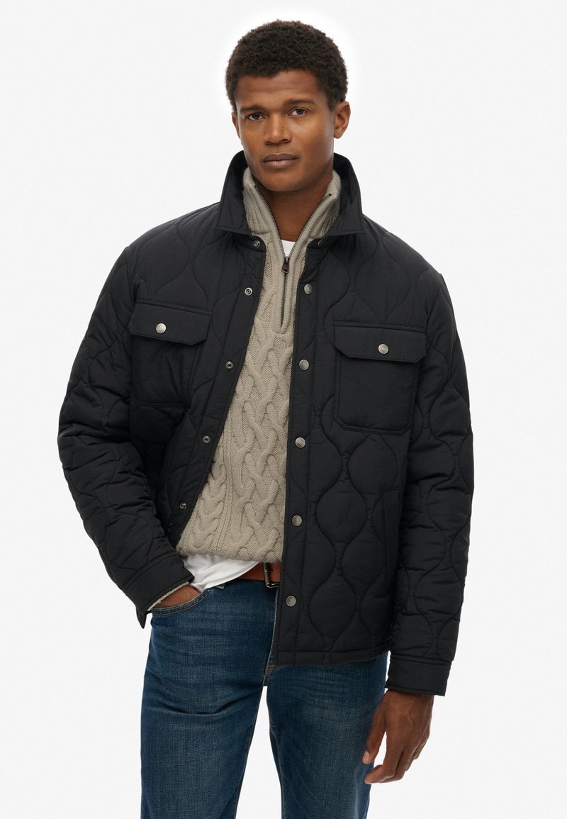 Schwarze gesteppte Jacke mit hohem Kragen, zwei Brusttaschen und Druckknöpfen, kombiniert mit einem cremefarbenen Strickpullover mit Zopfmuster.