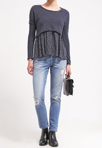 Marinblå krokad tröja över en blommig peplumtop, kombinerad med ljusblå slitna jeans och svarta ankelboots, hållande en svart handväska.