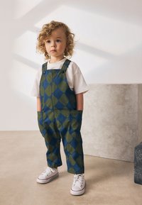 Salopette à carreaux bleus et verts avec des poches, associée à un t-shirt blanc et des baskets blanches. Le tissu semble doux et résistant.