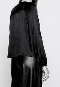 Donna in una blusa nera di satin a maniche lunghe abbinata a pantaloni in pelle nera a vita alta, in piedi contro uno sfondo neutro.