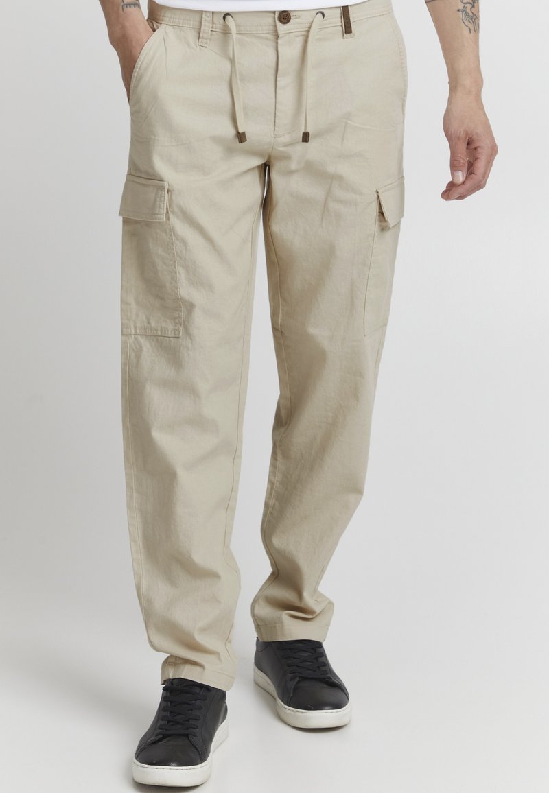 Pantalons cargo beiges avec une coupe décontractée, taille à cordon de serrage, poches avant et latérales, et détails de couture subtils, associés à des baskets noires.