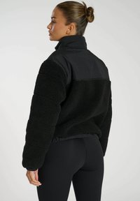 Schwarze Fleecejacke mit einem glatten schwarzen Oberteil, hohem Kragen, elastischem Saum und langen Ärmeln; verfügt über eine weiche, strukturierte Oberfläche.