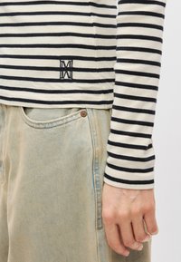 Ριγέ μακρυμάνικο πουκάμισο σε κρέμ και να�υτικό μπλε, με μικρό κεντημένο λογότυπο. Συνδυάζεται με ανοιχτό μπλε τζιν παντελόνι, που δείχνει λεπτομέρειες στις τσέπες.