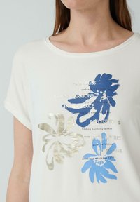 Witte T-shirt met korte mouwen, versierd met blauwe en gouden bloemenprints en tekst. Zacht materiaal; ronde halslijn; relaxte pasvorm.