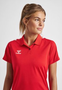 Rød polo shirt med krave, korte ærmer, og en hvid logo samt sorte knapper, glat tekstur og hvide detaljer på skuldrene.