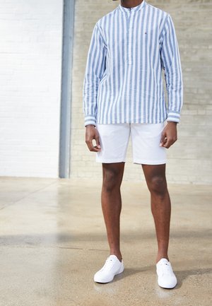 Homme portant une chemise à manches longues à rayures verticales bleues et blanches, un short blanc et des baskets blanches, debout à l'intérieur sur un sol poli.