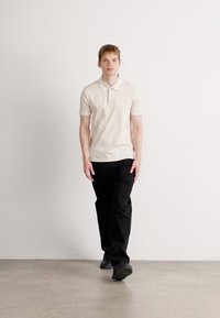 Modelul poartă un tricou polo bej deschis cu guler și mâneci scurte, asortat cu pantaloni negri și pantofi negri, stând în fața unui fundal simplu.