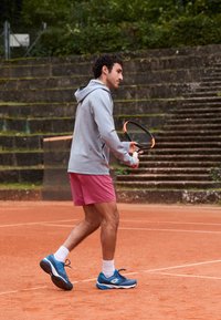Moški športnik v svetlo sivi kapucinskih jopiči in roza kratkih hlačah, drži teniško lopar na peščenem igrišču s kamnitimi stopnicami v ozadju.