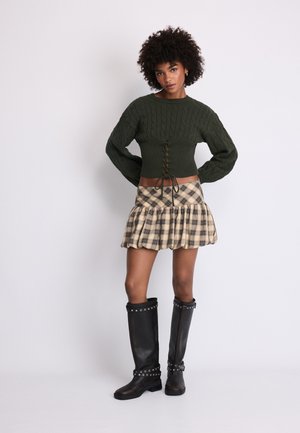 Donna con capelli ricci che indossa un maglione verde scuro lavorato a maglia, una minigonna beige a quadri con volant e stivali neri al ginocchio con cinturini e borchie, in piedi.