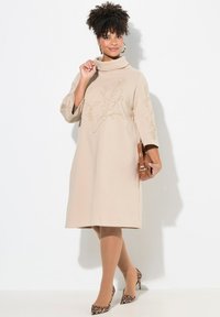 Beiges knielanges Kleid mit einem Cowl-Neck und drei Viertelärmeln, das mit floralem Stickereien verziert ist. Kombiniert mit High Heels im Leopardenmuster und einer Handtasche.