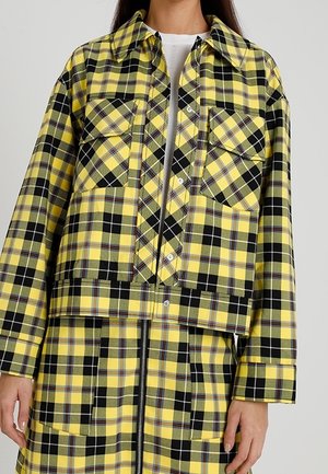 Kvinde iført en gul og sort ternet jakke med lommer foran og overlægsflap over et matchende ternet skørt med lynlås foran.