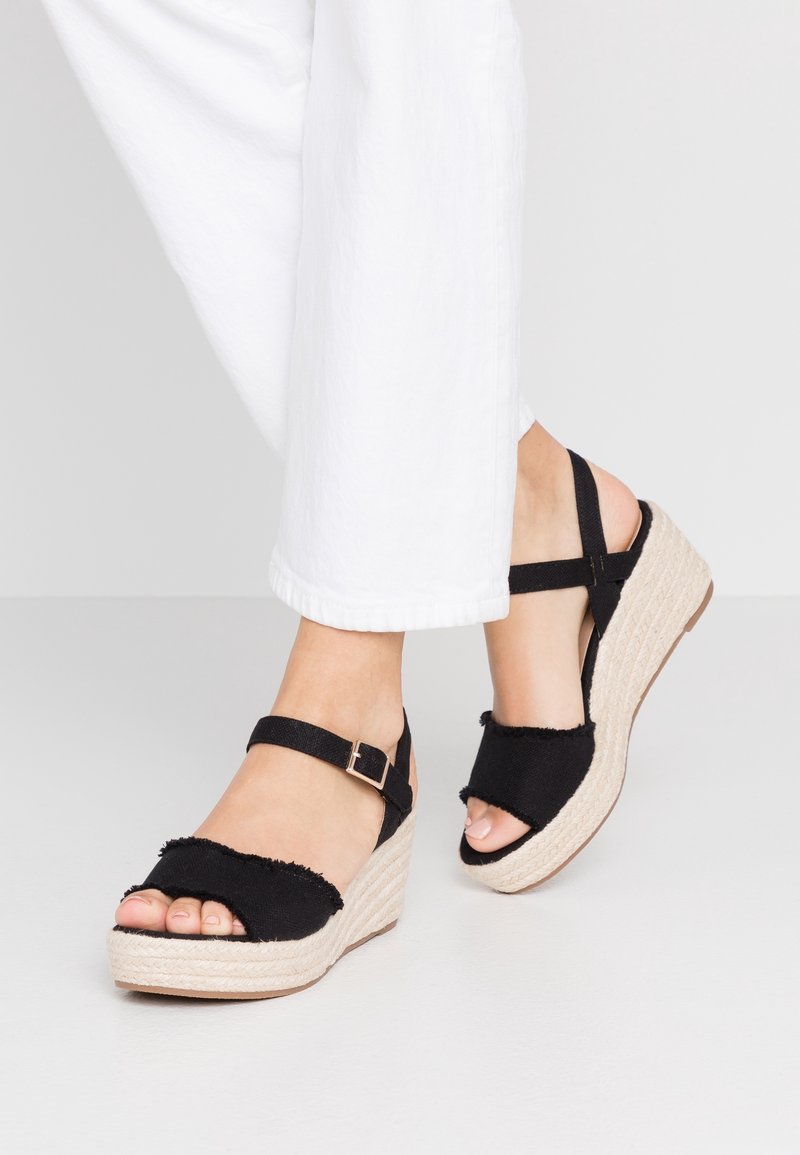 Pieds portant des sandales compensées en tissu noir avec des brides chevilles, assorties à un pantalon blanc court sur un fond clair uni.