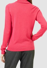 Pull à col roulé rose en matière douce, avec manches longues et ourlet côtelé. Porté avec un pantalon gris cintré, montrant une coupe ajustée.