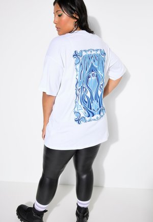 T-shirt oversize blanc avec un design floral abstrait bleu au dos. Associé à des leggings noirs brillants et des bottes à plateforme sombres.