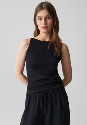 Jeune femme aux longs cheveux bruns portant un haut noir sans manches et un pantalon noir à cordon sur un fond gris uni.