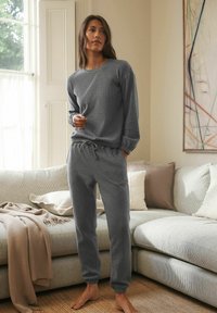 Grijze gebreide sweatshirt en bijpassende sweatpants met elastische boorden en een trekkoord in de taille, tegen een lichtkleurige bank en neutraal decor.