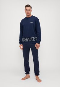 Set trenirki u tamnoplavoj boji Moschino, s logotipom na prsima i velikim podebljanim slovima duž nogavice. Mekana tkanina, ležeran kroj.