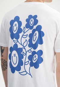 Witte T-shirt met een blauw bloemontwerp en lachende gezichten. De bloemen zijn gestileerd, met gebogen bloemblaadjes en groene stelen.