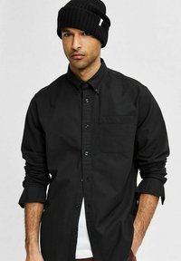 Camisa negra de botones con mangas largas, un bolsillo en el pecho y botones oscuros. Llevada con una gorra de punto negra. Textura de tela suave.