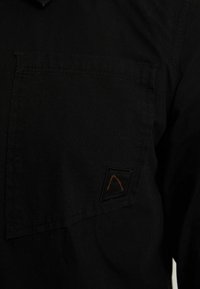 Zwarte katoenen shirt met een linkerkant borstzak, voorzien van een vierkante patch met een oranje bergontwerp. Soepele textuur, eenvoudig ontwerp.