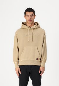 Felpa beige con cappuccio regolabile, tasca frontale e ricamo con la scritta "TOMMY JEANS". Polsini e orlo a coste; presenta una piccola etichetta con logo.