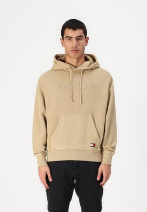 Beige hoodie with drawstring hood, front pouch pocket, and embroidered "TOMMY JEANS" text. Ribbed cuffs and hem; features a small logo patch.