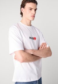 Balts kokvilnas T-krekls ar melnu tekstu "HUG" un sarkanu sirds grafiku, īsām piedurknēm, brīvu piegriezumu, gludu tekstūru.