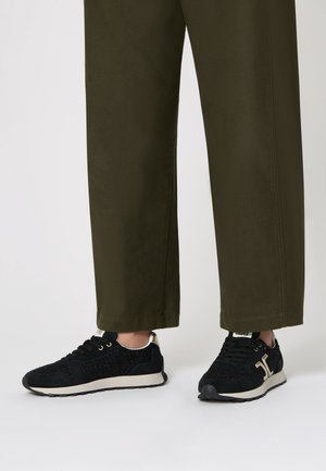 Sneakers neri con tessuto testurizzato e accenti in suede, dotati di una suola bianca e dettagli hardware dorati. Abbinati a pantaloni larghi verde oliva.