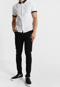 Camicia bianca a maniche corte con colletto nero, abbinata a pantaloni neri slim-fit e scarpe nere slip-on. Texture semplice.