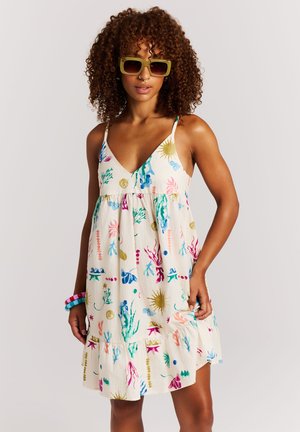 Vrouw met krullend haar die groene zonnebrillen draagt, een mouwloze witte jurk met kleurrijke koraal- en zonpatronen, en de zoom van de jurk vasthoudt.