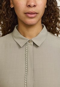 Gai pelēks blūze ar klasisku apkakli, ar pogu priekšpusi un dekoratīvu viļņotu apdari gar pogu līniju. Mīksta, teksturēta auduma.