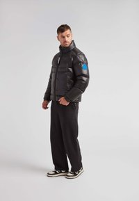 Sort pufferjakke med høj krave og lynlås, med quiltet tekstur og et blåt logo på venstre ærme. Sorte bukser og sneakers.