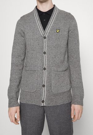 Homme portant un cardigan gris boutonné avec un liseré blanc, deux poches avant, un logo d'oiseau jaune et un pantalon gris à carreaux.