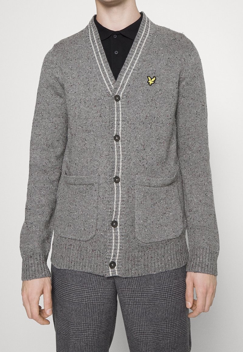 Homme portant un cardigan gris boutonné avec un liseré blanc, deux poches avant, un logo d'oiseau jaune et un pantalon gris à carreaux.