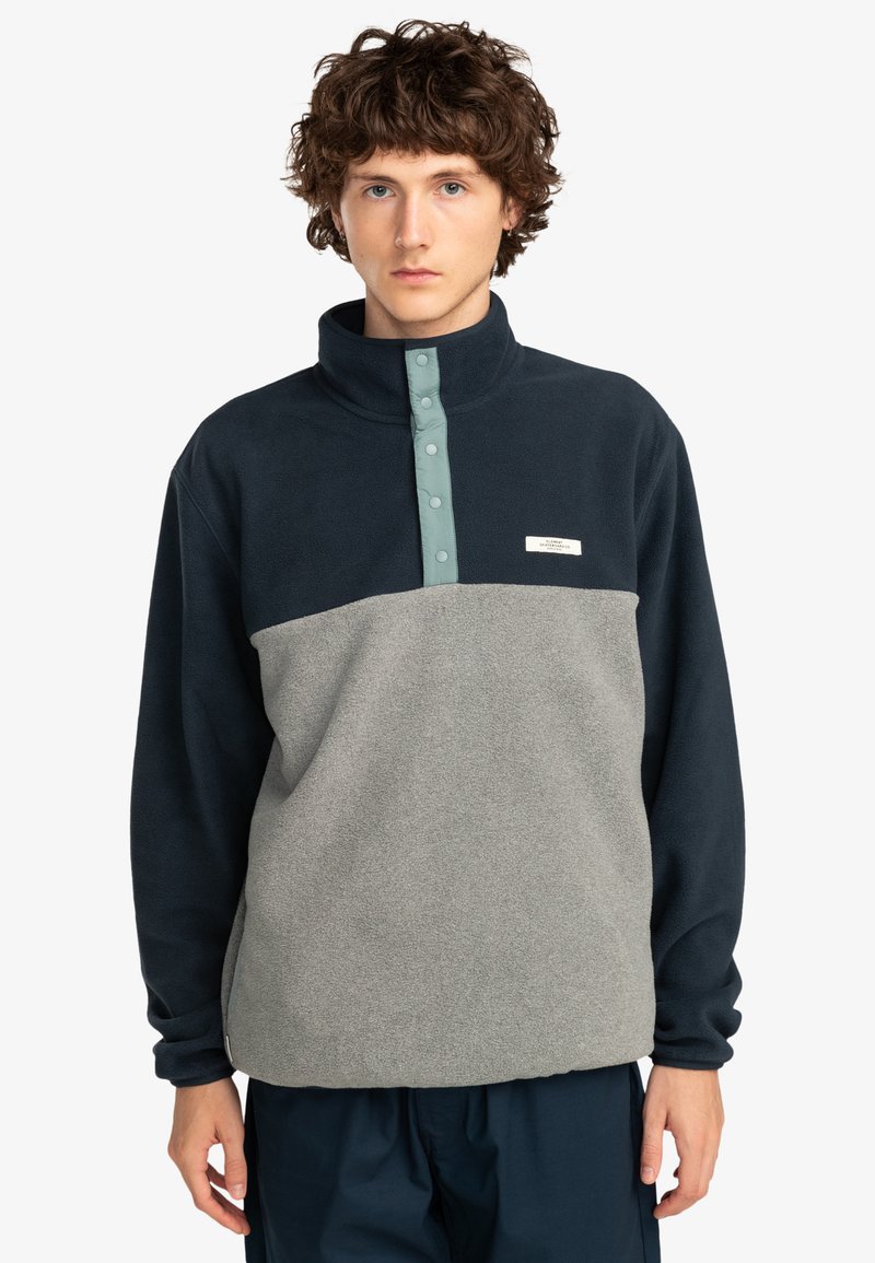 Fleece-pullover i mörk marinblått och ljusgrått med hög krage, tryckknappsplacket och en liten varumärkesetikett på bröstet. Mjuk textur.