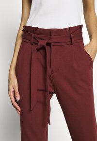 Maroon high-waisted pants with a tie belt, tapered legs, two side pockets, and a smooth texture. Plain design with no visible patterns.