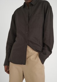 Dark brown button-up shirt made of textured fabric with a left chest pocket, worn tucked into beige trousers.